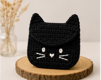 May include: A black crocheted pouch shaped like a cat head with pointed ears. The front features white embroidered details including two circular eyes, a small heart-shaped nose, and three whiskers on each side of the face, creating a feline face.