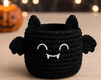 May include: A black crocheted basket shaped like a bat, featuring two pointed ears on top, two small wings on the sides, and a white embroidered face with closed eyes and two sharp fangs centered on the front of the round container.