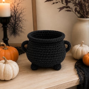 May include: A black crocheted cauldron with a wide circular opening, two small side handles, and three short rounded feet. The vessel is constructed from thick yarn with a visible textured stitch pattern, creating a deep, hollow interior space.