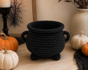 May include: A black crocheted cauldron with a wide circular opening, two small side handles, and three short rounded feet. The vessel is constructed from thick yarn with a visible textured stitch pattern, creating a deep, hollow interior space.