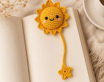 May include: A yellow crocheted sun bookmark with a smiling face, black bead eyes, and pink stitched cheeks. The sun has a long crocheted tail ending in a small yellow crocheted star, resting on the open pages of a blank white book.