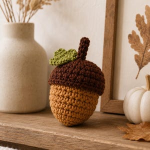 May include: A crocheted acorn ornament featuring a tan nut base, a dark brown textured cap, and a small green leaf attached to the top. The yarn is worked in a tight stitch pattern, creating a rounded shape with a short stem extending from the center.