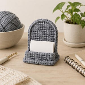 May include: A gray crocheted card holder with an arched back and a rectangular base, featuring a stack of blank white rectangular cards tucked into the front slot. The item is constructed from textured yarn stitches in a consistent gray color.