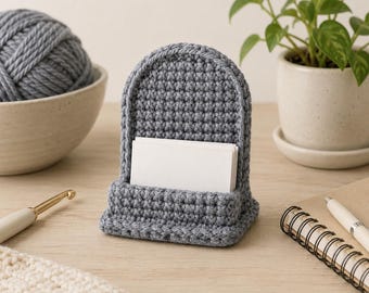 Pode incluir: Um porta-cartões de crochê cinza com encosto arqueado e base retangular, apresentando uma pilha de cartões retangulares brancos em branco inseridos na ranhura frontal. O item é feito de pontos de fio texturizados na cor cinza.