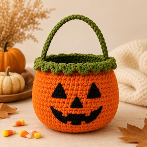 May include: A round, orange crochet basket featuring a jack-o'-lantern face with black yarn eyes, a nose, and a wide, jagged mouth. The basket has a green crocheted rim with a scalloped edge and a matching green braided handle attached to the top.