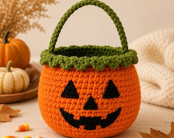 Op de afbeelding: Een ronde, oranje gehaakte mand met een jack-o'-lantern gezicht van zwart garen, bestaande uit ogen, een neus en een brede, grillige mond. De mand heeft een groene gehaakte rand met een schulprand en een bijpassend gevlochten handvat.