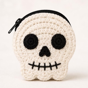 Puede incluir: Un monedero redondo de ganchillo en color blanco roto con forma de calavera. La calavera presenta dos ojos circulares de ganchillo negro, una pequeña nariz triangular negra y una boca negra bordada con líneas verticales. Una cremallera negra está fijada en la parte superior.