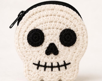 May include: A round, off-white crocheted coin purse shaped like a skull. The skull features two circular black crocheted eyes, a small black triangular nose, and a stitched black mouth with vertical lines. A black zipper is attached at the top.