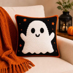 Puede incluir: Un cojín cuadrado de ganchillo con fondo negro y un gran fantasma blanco en el centro. El fantasma tiene dos ojos ovalados negros y una pequeña boca curva. Pequeñas estrellas y puntos de color naranja están dispersos, con un borde de color naranja.