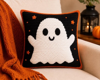 May include: A square crochet pillow featuring a black background with a large white ghost in the center. The ghost has two black oval eyes and a small curved mouth. Small orange stars and dots are scattered around the ghost, and the pillow has an orange border.