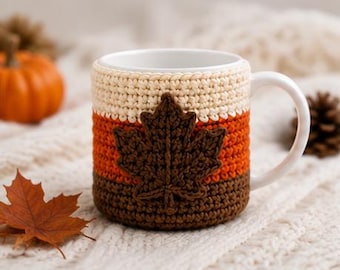 May include: A white ceramic mug featuring a removable crochet cozy. The cozy is designed with three horizontal color bands in cream, orange, and brown, and it is decorated with a prominent dark brown crocheted maple leaf on the front side.