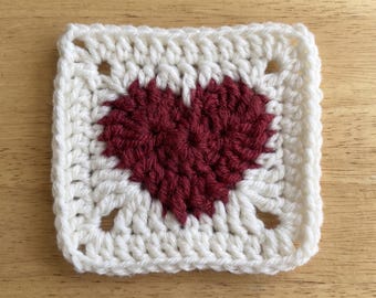 May include: A square crochet motif featuring a dark red heart centred within a white border. The piece is constructed with double crochet stitches, creating a textured pattern that transitions from the heart shape to a square frame.