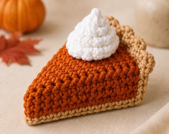 May include: A crochet slice of pumpkin pie featuring a deep orange filling, a light tan crust with a scalloped edge, and a dollop of white whipped cream on top. The piece is crafted with visible yarn stitches and has a triangular shape.