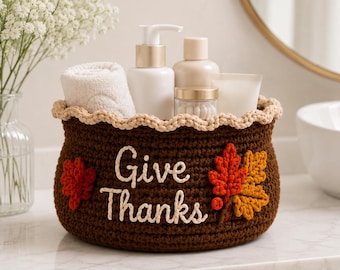 May include: A dark brown crocheted basket with a beige scalloped rim, featuring the white embroidered text "Give Thanks" and decorative red and orange crocheted autumn leaves on the front, holding a white towel and various toiletry bottles.