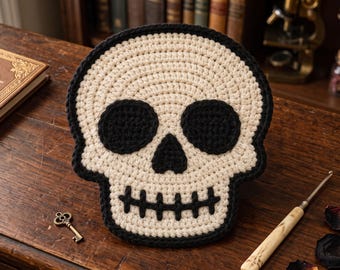 May include: A crochet skull wall hanging or decor piece featuring a cream-colored base with black yarn detailing for the eye sockets, nose cavity, and stitched mouth. The skull is outlined in a thick black border, creating a high-contrast design.