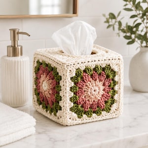 May include: A square tissue box cover featuring a crochet granny square pattern in cream, olive green, and dusty rose yarn. The cover is constructed with a thick, textured yarn and has an open top to allow white facial tissues to be pulled through.
