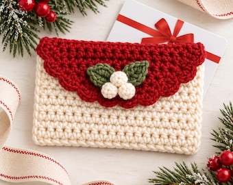 May include: A rectangular cream-colored crochet pouch featuring a scalloped red flap adorned with two green crocheted leaves and three small white crocheted berries, designed to hold a gift card or small items.