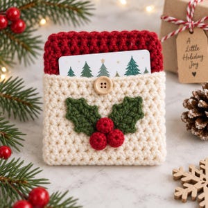 Puede incluir: Un tarjetero de ganchillo color crema con un borde superior rojo, cierre de botón de madera y una aplicación decorativa de hojas de acebo verdes y bayas rojas. Una tarjeta blanca con un patrón de árboles de Navidad está insertada.