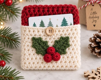 May include: A cream-colored crochet gift card holder featuring a red crochet top border, a wooden button closure, and a decorative green holly leaf and red berry applique. A white card with a green Christmas tree pattern is tucked inside.