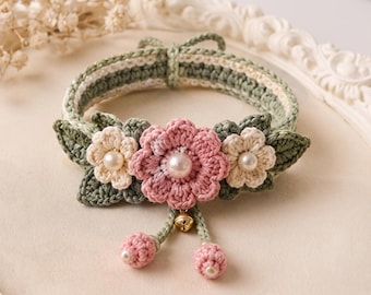 May include: A crocheted collar featuring a sage green band with white stripes, adorned with a central pink flower, two smaller cream flowers, and green leaves. Each flower has a central pearl bead. Two pink crocheted spheres and a small gold bell hang below.