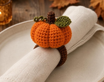 May include: A crochet napkin ring featuring a bright orange pumpkin with a dark brown stem, a small green leaf, and a curly green vine. The pumpkin is attached to a solid dark brown circular band that holds a folded off-white linen napkin.
