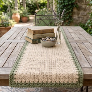 May include: A rectangular crochet table runner featuring a cream-colored center with a sage green border. The runner is placed on a wooden table, topped with a stack of three books, a small bowl filled with smooth stones, and a glass vase with lavender.