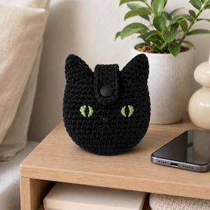 Puede incluir: Un estuche de ganchillo negro con forma de cabeza de gato, orejas puntiagudas, dos ojos bordados en verde lima y un cierre central de botón a presión negro. Este pequeño accesorio textil redondo y texturizado tiene una nariz bordada.
