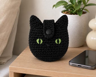 May include: A black crocheted pouch shaped like a cat head with pointed ears, two embroidered lime green eyes, and a central black snap button closure. The small, round, textured textile accessory features a simple stitched nose detail.