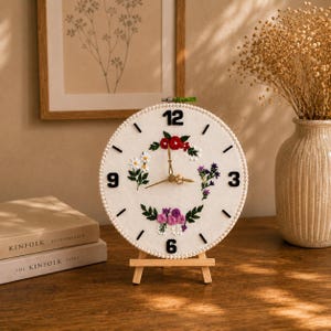 May include: A round wall clock featuring a white fabric face with embroidered floral clusters in red, white, purple, and pink. The clock has black numerals at 3, 6, 9, and 12, black tick marks for other hours, gold hands, and a white beaded border.