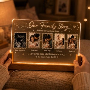 May include: A rectangular acrylic light panel set in a wooden base, featuring the text "Our Family Story" and "From the first heartbeat to forever." It displays five photo frames showing an ultrasound and family moments, with "The Bennett Family · Est. 2019" at the bottom.