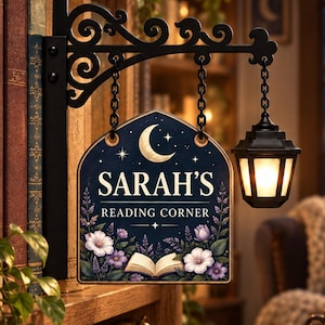 May include: A decorative hanging sign with a black metal bracket and a small lantern. The sign features a dark blue background with a crescent moon, white flowers, an open book, and the text "SARAH'S READING CORNER" in white lettering.