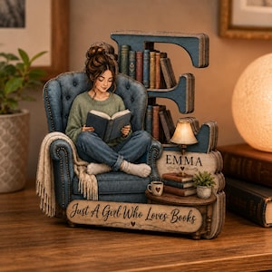 May include: A decorative wooden letter E featuring a figure sitting in a blue tufted armchair reading a book. The piece includes miniature books, a lamp, a small table, and the text "EMMA" and "Just A Girl Who Loves Books" printed on the base.