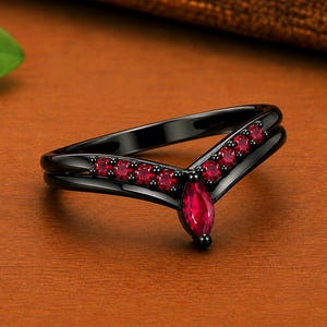 May include: A V-shaped ring crafted from black metal, featuring a central marquise-cut red gemstone at the point, flanked by a series of smaller round-cut red stones set along both sides of the band in a symmetrical arrangement.