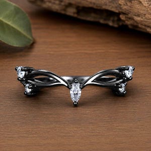 Könnte beinhalten: Ein Ring aus schwarzem Metall mit einem geschwungenen, V-förmigen Banddesign. Der Ring ist mit fünf klaren, farblosen Steinen besetzt: einem zentralen Stein im Marquise-Schliff an der Spitze und zwei kleineren Rundschliff-Steinen an den Armen.