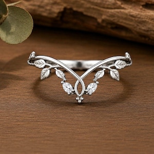 May include: A silver-toned V-shaped ring featuring a delicate vine motif with engraved leaves and two marquise-cut clear stones set at the centre point. The band has a polished finish and a symmetrical design that curves upwards to a central peak.