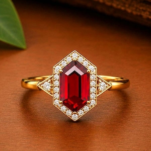 Könnte beinhalten: Ein Goldring mit einem zentralen hexagonalen roten Edelstein, der von einem Kranz aus kleinen runden weißen Steinen umgeben ist. Die Fassung enthält auf jeder Seite dreieckige Akzente, die ebenfalls mit kleinen weißen Steinen besetzt sind.