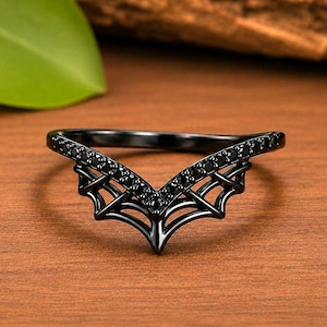 May include: A black ring featuring a V-shaped band with a web-like design underneath the top section. The upper portion of the band is encrusted with small, dark, circular stones that follow the chevron contour of the ring's structure.