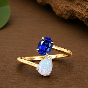 Könnte beinhalten: Ein goldener Bypass-Ring mit zwei verschiedenen Edelsteinen. Ein Ende des Rings trägt einen leuchtend blauen Saphir im Ovalschliff, während das andere Ende einen tropfenförmigen, schillernden weißen Opal auf einer polierten Goldschiene zeigt.