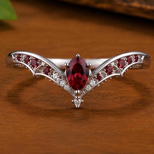 Puede incluir: Un anillo de plata en forma de V con una piedra preciosa de color rojo intenso en talla marquesa en el centro. La banda está decorada con piedras rojas redondas más pequeñas y diminutas piedras blancas transparentes a lo largo del marco.