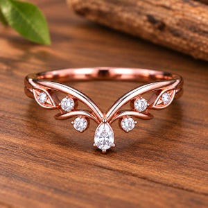 Könnte beinhalten: Ein V-förmiger Ring aus Roségold mit einem zentralen, birnenförmigen, klaren Edelstein und sechs kleineren, runden, klaren Edelsteinen entlang der Ringschiene. Das Metall ist poliert und das Design hat eine geschwungene Chevron-Form.