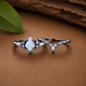 Könnte beinhalten: Ein zweiteiliges Ring-Set aus schwarzem Metall mit einem zentralen drachenförmigen Opal, akzentuiert durch kleine runde violette Steine und marquiseförmige Opale. Der zweite Ring ist ein V-förmiges Band mit einer Reihe kleiner violetter Steine.