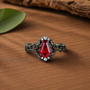 Könnte beinhalten: Ein schwarzer Ring mit einem großen, roten, sargförmigen Edelstein in der Mitte. Der Ring ist mit kleinen weißen und roten Edelsteinen und floralen Details verziert. Der Ring liegt auf einer Holzoberfläche.