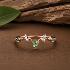 Könnte beinhalten: Ein Ring aus Roségold mit einem zentralen, marquise-geschliffenen grünen Rutilquarz, flankiert von zwei kleinen runden grünen Steinen und vier klaren runden Akzentsteinen in einer zarten, symmetrischen, rankenartigen Metallfassung.