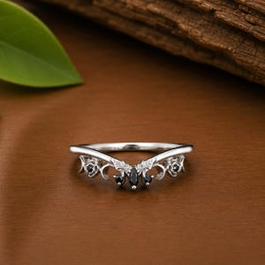 May include: A silver-toned ring with a curved design, featuring black gemstones at the center and decorative floral and scrollwork accents. The ring is set against a brown wooden surface.