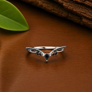 May include: A silver ring with a V-shaped design. It has a central black gemstone and smaller black stones along the curved edges. The ring is set against a warm brown background. The ring is likely made of silver.