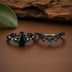 Könnte beinhalten: Ein Ring-Set aus schwarzem Metall mit einem zentralen sargförmigen schwarzen Stein, flankiert von Fledermausflügel-Motiven mit kleinen klaren Steinen, sowie einem passenden V-förmigen Chevron-Ring, der mit winzigen schwarzen Steinen besetzt ist.