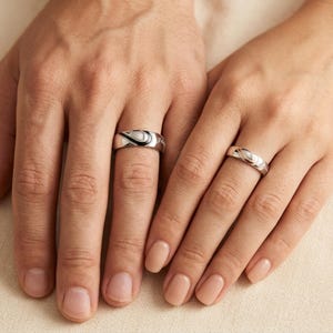 May include: Two silver-toned metal rings worn on fingers. Each ring features a polished band with a central inlay design that resembles a stylized infinity symbol or a curved wave pattern, creating a cohesive look across both pieces of jewelry.