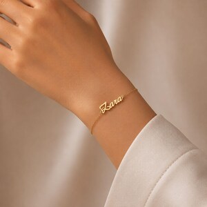 Op de afbeelding: Een delicate gouden armband met de naam "Zara" in cursief schrift. De armband wordt om een pols gedragen, waarbij het elegante ontwerp en de fijne details van de gepersonaliseerde sieraden worden getoond. De armband is een eenvoudige ketting met de naam als middelpunt.