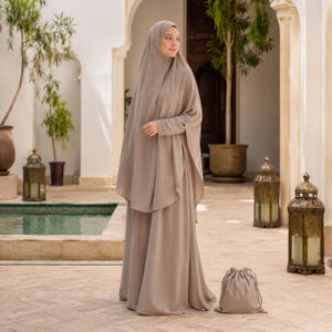 May include: A solid taupe-coloured prayer dress featuring a long, flowing skirt and an attached head covering that drapes over the shoulders and torso. The garment includes long sleeves and a matching drawstring storage pouch placed on the ground.