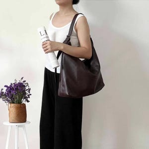 May include: A large, dark brown hobo-style shoulder bag with a smooth, matte finish. The bag features a single wide strap with an adjustable metal buckle, a slouchy, unstructured silhouette, and a spacious main compartment with a curved top opening.